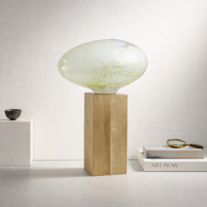 Designerska lampa ze szkła artystycznego na drewnianym cokole, sfotografowana pod kątem w eleganckiej, jasnej przestrzeni, z subtelnymi dodatkami podkreślającymi skalę.